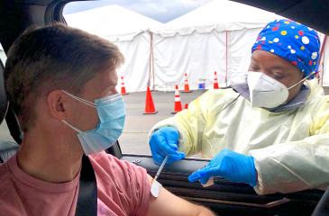 A nurse in scrubs, a mask, and gloves administers a vaccine to a masked person in their vehicle